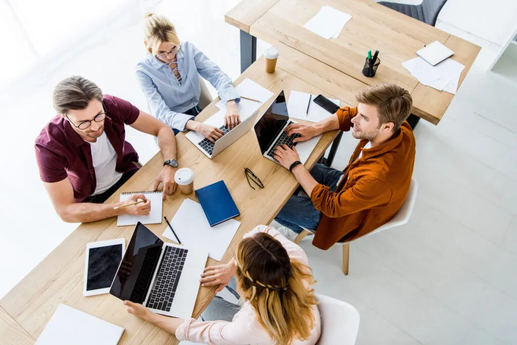 Four professionals collaborate at a wooden table with laptops, notebooks, and coffee cups in a bright office setting.
