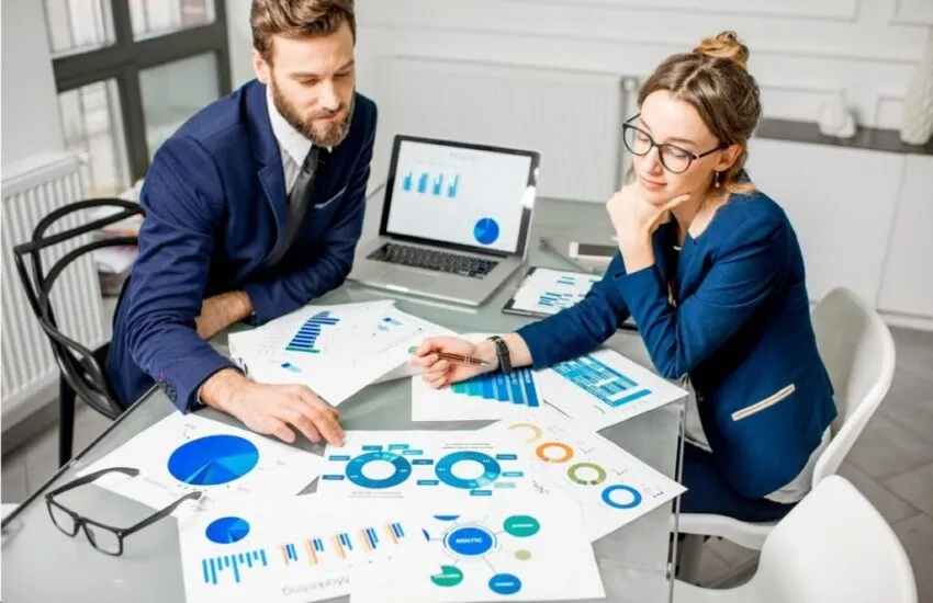 Two professionals discuss marketing data charts and graphs spread on a table with a laptop showing analytics.